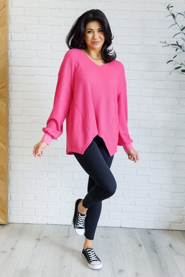 A woman is posing in a pink V-neck top with banded sleeve cuffs and a scooped asymmetrical hemline, standing in a room with white brick walls and dancing in a pair of black sneakers.