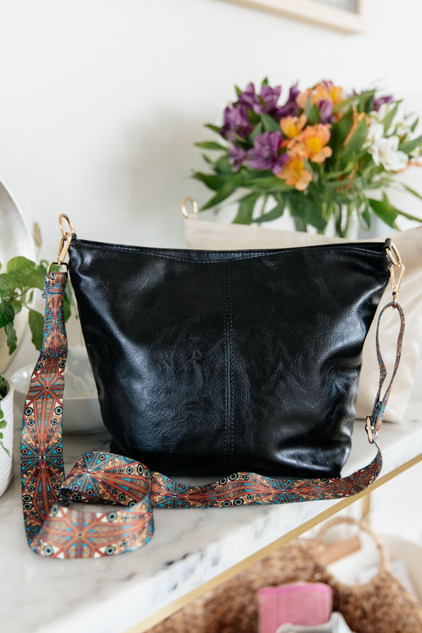 Black leather handbag with a colorful strap on a white surface with flowers in the background