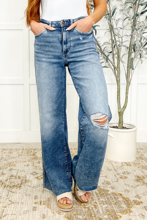 A pair of high-waisted, vintage wash blue denim wide-leg jeans with distressed knees and a raw hem, worn by a model.